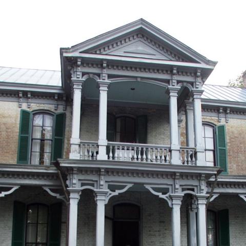 Two-story brick house with green shutters, ornate white columns, and a central balcony under a triangular pediment.